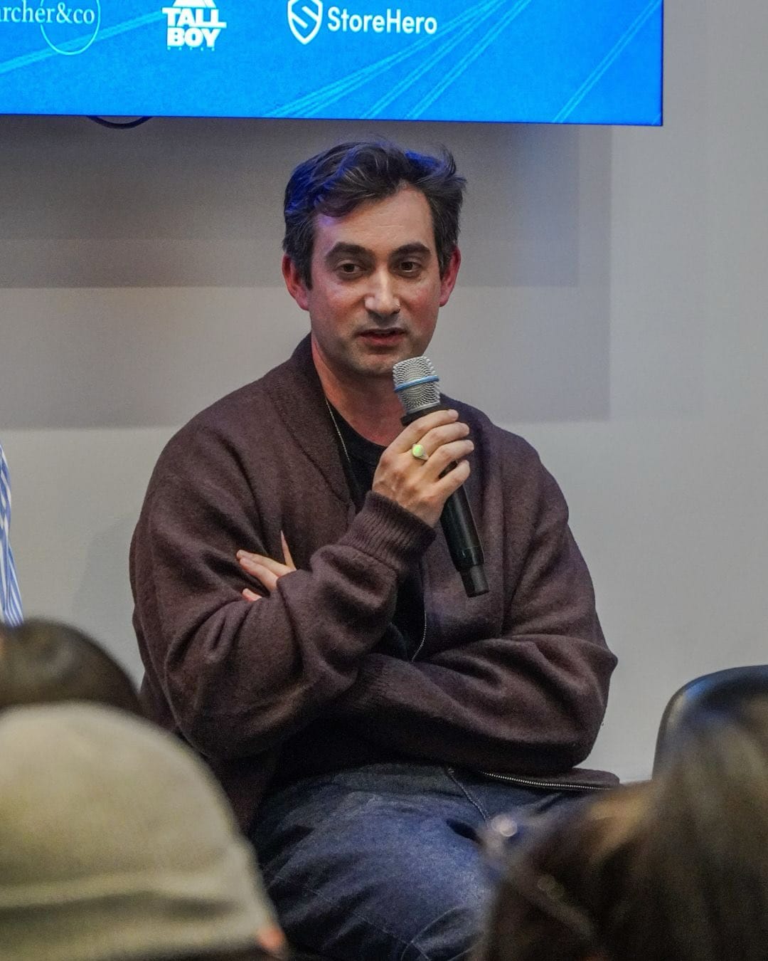 A panelist in glasses and a dark blazer speaks into a handheld microphone, with the Shopify logo on the screen behind him.